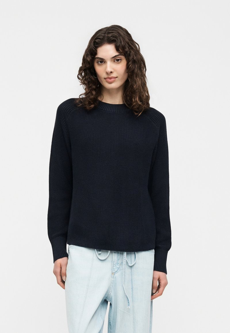 Femme aux cheveux bruns bouclés portant un pull en maille noir et un jean taille haute bleu clair, debout devant un fond blanc uni.