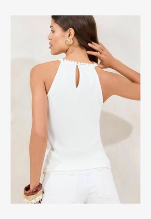White sleeveless top with scalloped trim, keyhole back detail, and ribbed texture. Paired with a white bottom and multi-colored bangles.