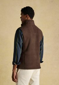 Hnedá fleecová vesta má vysoký golier, logo vyrazené na chrbte a bočné vrecká, skombinovaná s modrou pruhovanou košeľou a svetlými nohavicami.