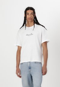 T-shirt bianca in cotone a maniche corte, con il logo "Premium Frost" in corsivo nero sul petto, abbinata a jeans azzurri chiari.