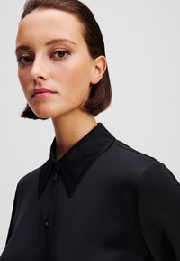 Camicia nera con colletto, dalla texture liscia, caratterizzata da un'apertura con bottoni e un luminoso sottotono. Il colletto presenta un dettaglio di bordatura in contrasto.