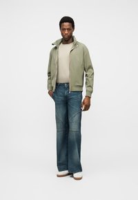 Νεαρός άνδρας που φοράει ανοιχτοπράσινο μπουφάν με φερμουάρ, μπεζ μπλουζάκι, φαρδιά μπλε τζιν και λευκά αθλητικά παπούτσια, στέκεται μπροστά σε απλό λευκό φόντο.