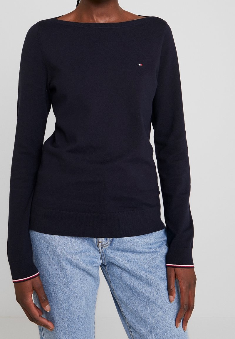 Pull en maille bleu marine avec manches longues, encolure large et poignets côtelés arborant des rayures rouges et blanches à l'ourlet. Détail minimaliste du logo présent.