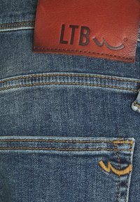 Takapocket farku denimissa, jossa on oranssi tikkaus ja ruskea nahkalaatta, jossa lukee "LTB" vyötäröllä.