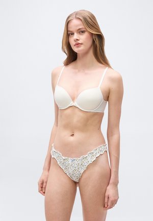Jeune femme aux longs cheveux blonds portant un soutien-gorge blanc rembourré et un slip bikini à volants fleuris, debout devant un fond uni.