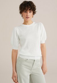 Top corto a maniche corte in maglia bianca con maniche a palloncino, scollatura rotonda, orlo a coste, indossato con pantaloni di colore chiaro, caratterizzati da una consistenza liscia.
