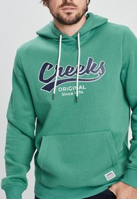 Sudadera con capucha verde en tejido suave, que presenta un gran texto gráfico en blanco y marino "Creeks Original Desde 1974" y un bolsillo frontal.