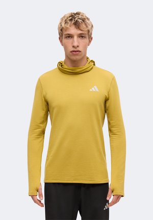 Jeune homme aux cheveux blonds courts portant un sweat à capuche de sport jaune moutarde à manches longues et un pantalon Adidas noir, debout devant un fond uni.