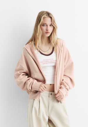 Helle rosa Zip-Hoodie mit weicher Textur, lockerem Schnitt und Kängurutaschen. Getragen über einem weißen cropped Top mit pinkem Text.