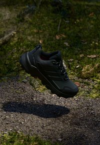 Schwarzer Sportschuh mit strukturiertem Obermaterial, auffälligem Schnürsystem und robustem Profil, schwebend über einem felsigen, natürlichen Hintergrund.
