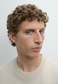 Portret osoby z kręconymi blond włosami, ubraną w beżową koszulkę z okrągłym dekoltem, na tle jasnoszarym. Minimalistyczne i neutralne tonacje.