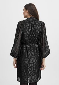 Rochie neagră cu un overlay transparent, mâneci bufante și un model de imprimeu animal pe întreaga suprafață. Include o talie legată și o textură ușoară.