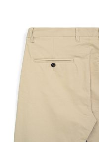 Beige chino pants featuring a back pocket with a black button closure. Smooth cotton fabric with a tailored waist and subtle stitched detailing.