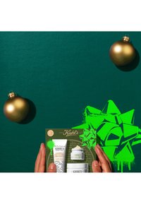 Mains tenant un coffret cadeau de soins de la peau Kiehl's pour les fêtes avec des ornements dorés et un nœud vert néon sur un fond texturé vert foncé.