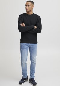 Schwarzer Ripp-Pullover mit Rundhalsausschnitt, kombiniert mit hellblauen distressed Jeans und schwarzen Laufschuhen. Das Model steht mit verschränkten Armen.