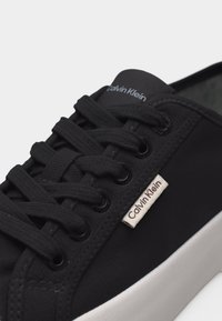Baskets Calvin Klein noires avec lacets en tissu, œillets en métal et semelle blanche, présentant une petite étiquette logo Calvin Klein sur le côté.