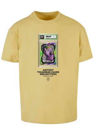 Gul bomulds t-shirt med en grafisk tryk af et samlekort med en lilla karakter. Teksten lyder "Artist Trading Card Selection."