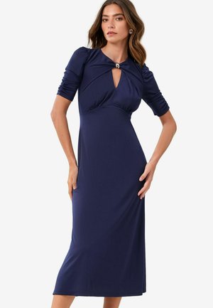 Navy blauwe jurk van rekbaar materiaal, met een sleutelgat-halslijn en korte pofmouwen. Strak om de taille, uitlopend naar een lengte tot halverwege de kuit.