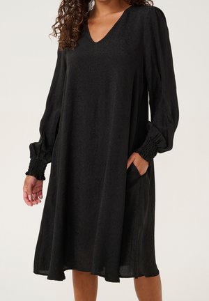 Žena v černých texturovaných midi šatech s dlouhými nabíranými rukávy a naškrobenými manžetami, rukou v kapse, stojící před jednobarevným pozadím.