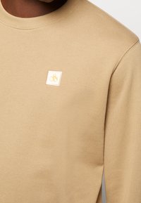 Maglione beige con collo rotondo e polsini a coste, con un piccolo logo giallo e bianco sul petto. Tessuto morbido.