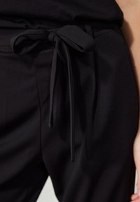 Pantalon noir avec une ceinture en tissu nouée en nœud à la taille, montrant un gros plan de l’avant et d’une partie de la main droite.