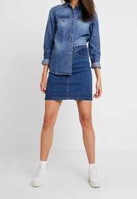 Jeanshemd- und Rock-Set in Blau. Das Hemd hat zwei Brusttaschen und eine Knopfverschluss. Der Rock hat ein tailliertes Design mit einer Vordernaht. Weiße Sneaker.