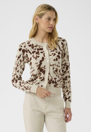 Beige neulejakku, jossa on ruskeita laikkuja, napit edessä ja ribbikäänteet hihansuissa ja helmassa. Pehmeä neulosmateriaali, joka sopii kerrospukeutumiseen.