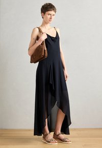Black asymmetrical dress in lightweight fabric, featuring a cowl neckline and embellished straps. Paired with a brown shoulder bag and leopard sandals.