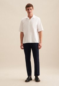 Witte kortarmige button-up shirt gecombineerd met navy lichtgewicht broeken en zwarte andermanschoenen, staand op een lichte achtergrond.