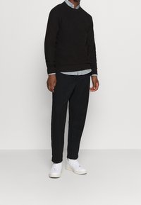 Tumma ruskea neulepusero, jonka alla vaaleansininen kauluspaita, yhdistetty mustiin kapealahkeisiin housuihin ja valkoisiin lenkkareihin. Yksinkertainen, moderni muotoilu.