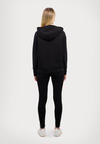 Felpa nera imbottita con cappuccio, abbinata a leggings neri. Presenta polsini e orlo a costine, indossata con sneaker bianche. Vista posteriore.