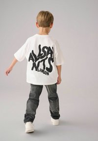 Bílý oversized tričko se stylovým černým nápisem "ALLSAINTS LONDON." Kombinováno s tmavě šedými džíny a bílými teniskami.