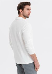 Man med brunt hår som bär en vit långärmad polo-shirt och grå rutiga byxor, tittar åt sidan mot en vit bakgrund.