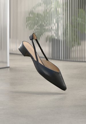 Svart lær slingback flatformet sko med spiss tå og lav blokkhæl, svevende over et betonggulv innendørs.