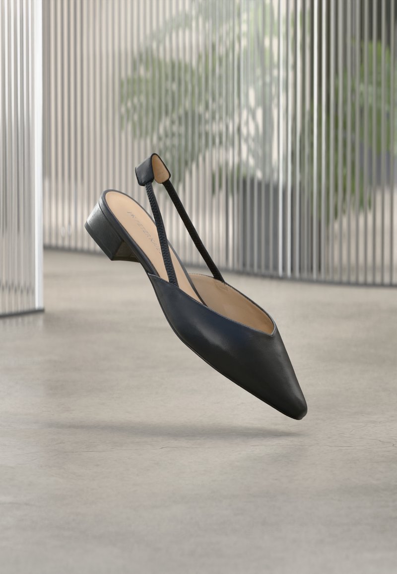 Μαύρο δερμάτινο γόβα slingback με μυτερή μύτη και χαμηλό χοντρό τακούνι που αιωρείται πάνω από δάπεδο από σκυρόδεμα εσωτερικού χώρου.