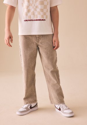 Person trägt beige Jeans mit geradem Bein, weiße Sneaker mit dunklem Logo und weißes T-Shirt mit beigem Schachbrettmuster und rotem Text.