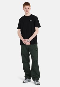 Zwarte katoenen t-shirt met korte mouwen. Olijfgroene cargobroek met zijzakken. Grijze hardloopschoenen met zwarte accenten.
