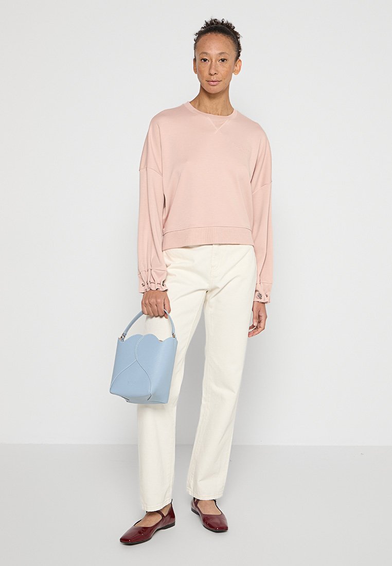 Helles rosa, übergroßes Sweatshirt mit Rundhalsausschnitt und gerafften Ärmeln, kombiniert mit weißen, geraden Hosen. Hält eine hellblaue strukturierte Handtasche.
