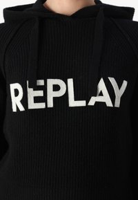 Haut à capuche en tricot noir avec une texture côtelée et un logo "REPLAY" en fil argenté brodé sur le devant. Dispose d'une capuche avec cordon de serrage.