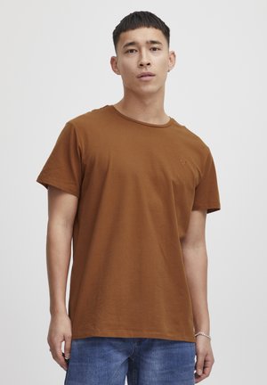 Hombre con el cabello corto que lleva una camiseta marrón lisa y vaqueros azules, de pie frente a un fondo gris claro.