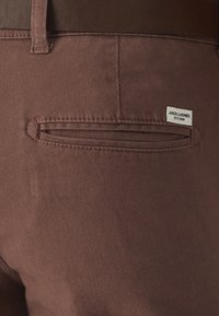 Braune Baumwollhose mit strukturierter Oberfläche, ausgestattet mit einer schräge Tasche und einem markierten Etikett, das auf der Rückseite aufgenäht ist.