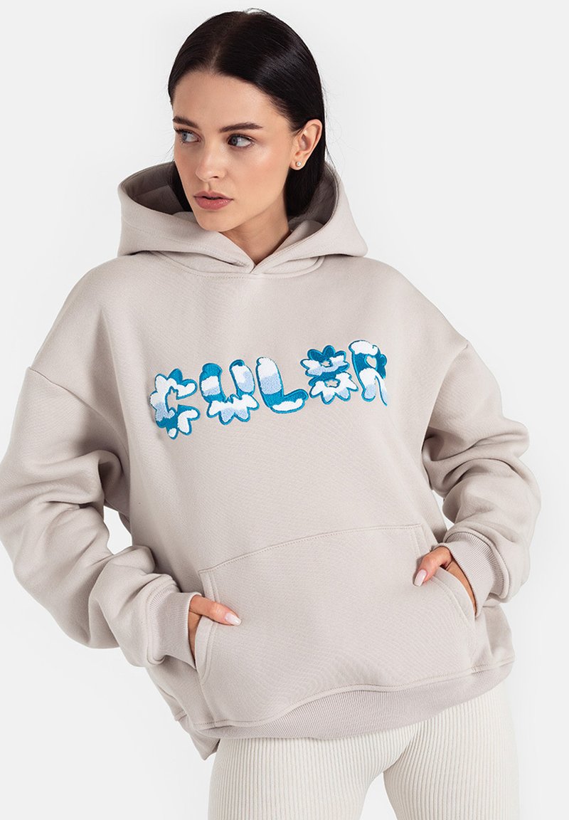 Beżowa bluza z kapturem z przednią kieszenią i odważnym niebieskim graficznym napisem w kwiaty. Miękki materiał z luźnym krojem i prążkowanymi mankietami.