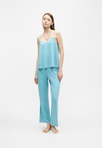 Jeune femme debout pieds nus portant un top bleu clair sans manches et un pantalon large assorti sur fond blanc.