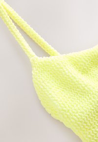 Neono geltonos spalvos bikini viršus su raukšlėtu tekstūros raštu. Turi plonas petnešėles ir išlenktą iškirptę, išryškinančią raukšlėto audinio dizainą.