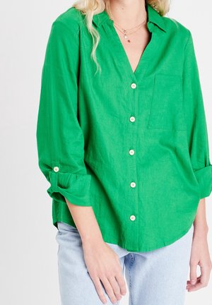 Personne portant une chemise boutonnée vert vif avec manches retroussées, associée à un jean bleu clair, ne montrant que le torse et les bras.