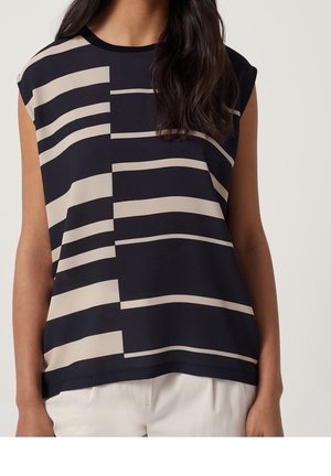 Ärmelloses schwarzes Top mit beigen horizontalen und vertikalen rechteckigen Streifen, getragen zu weißen Hosen, gezeigt von den Schultern bis zur Hüfte.