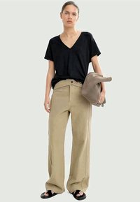 Fekete rövid ujjú V-nyakú póló, bézs oversize nadrág oldalsó varrás részletezéssel, és fekete szandál. A modell egy szürke táskát tart.