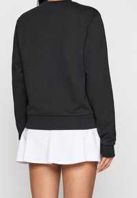Personne portant un haut noir à manches longues et une jupe plissée blanche courte avec de petites perforations, vue de dos.