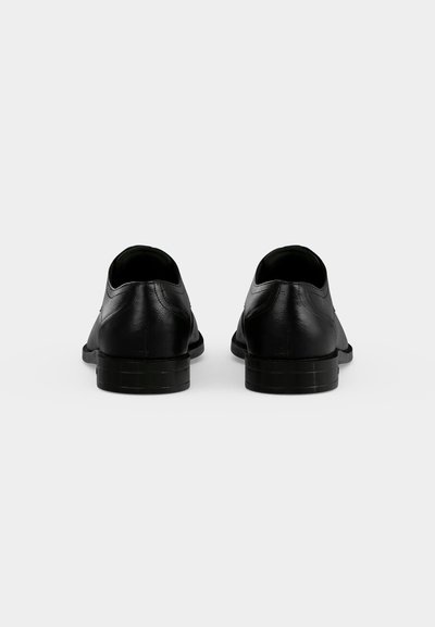 Paire de chaussures formelles en cuir noir vues de dos avec des talons bas sur fond blanc uni.