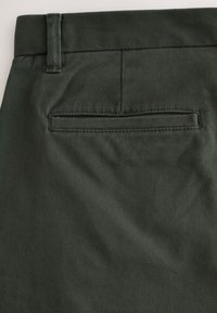 Gros plan sur la poche arrière d'un pantalon vert foncé avec passants de ceinture, présentant une poche passepoilée horizontale et des détails de coutures.